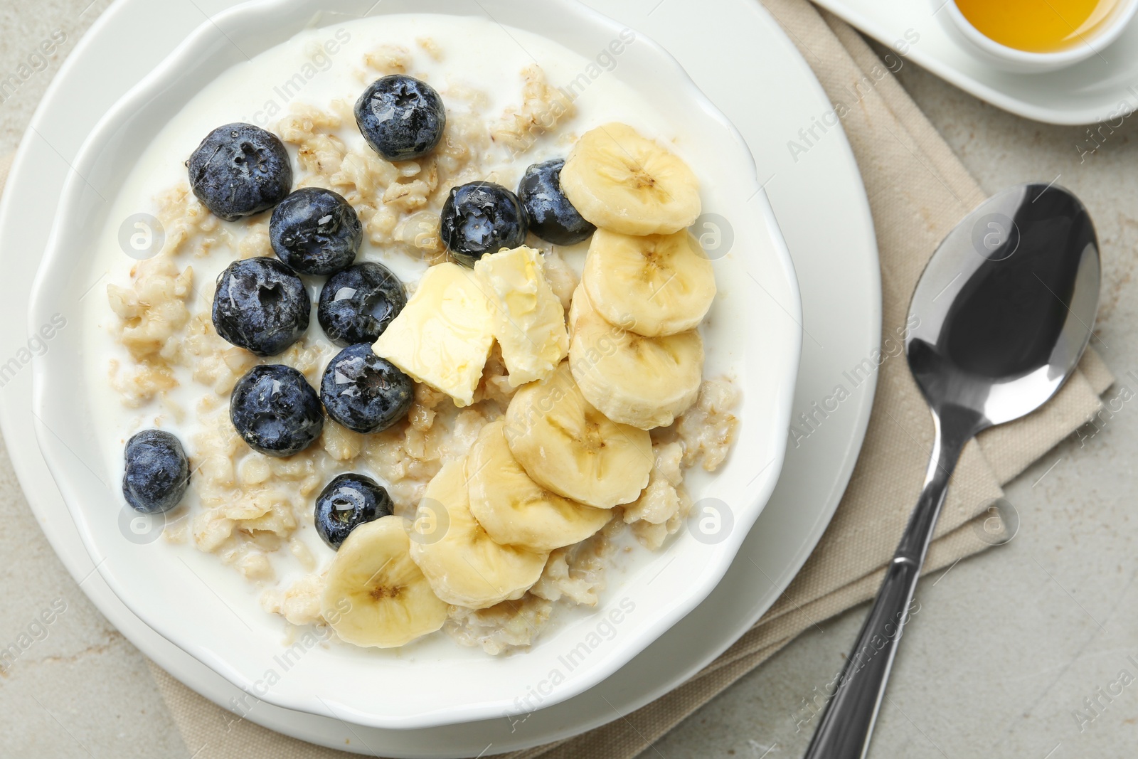 Photo of Tasty oatmeal with banana, blueberries, butter and milk served in bowl on light grey table, flat lay