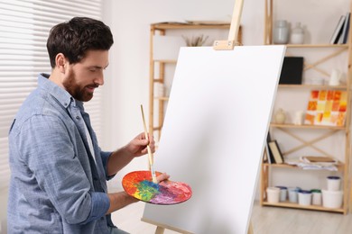 Man with brush and palette painting in studio. Using easel to hold canvas