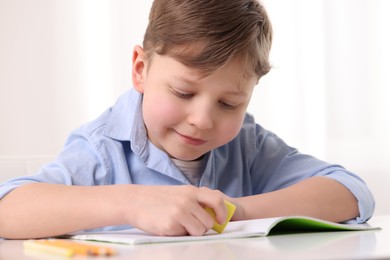 Photo of Little boy erasing mistake in his notebook at white desk indoors