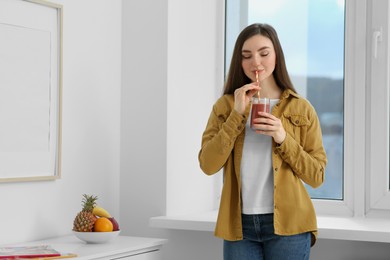 Photo of Beautiful young woman drinking delicious smoothie near window at home. Space for text