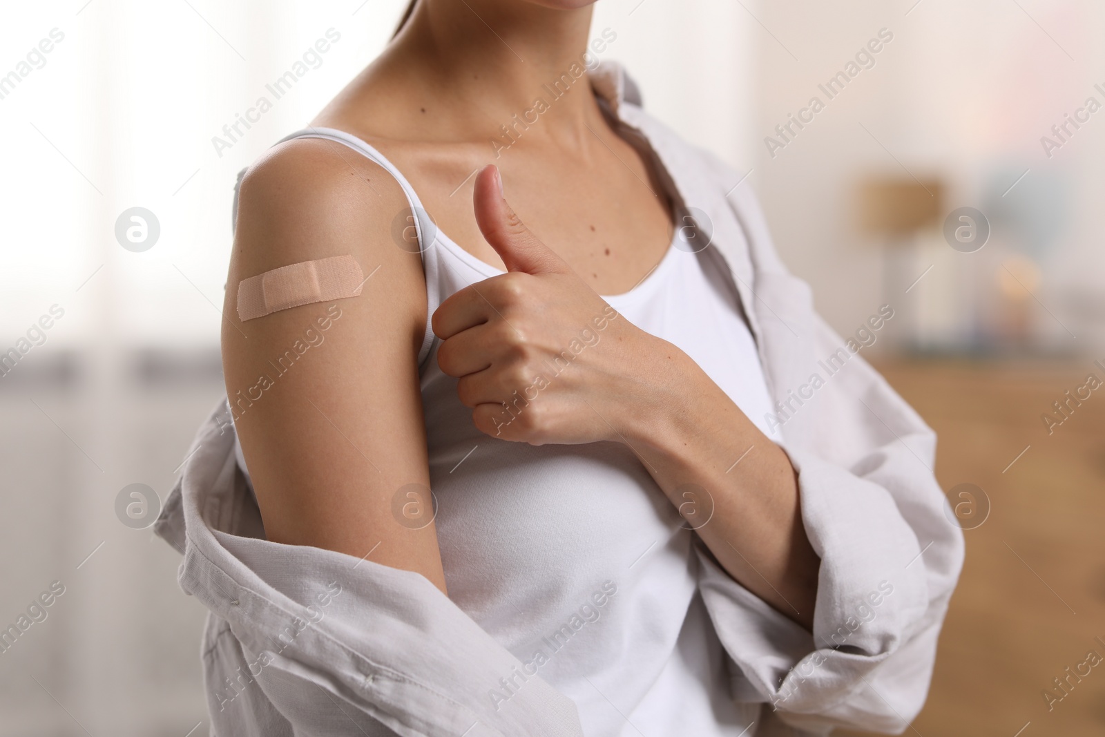 Photo of Woman with sticking plaster on arm after vaccination showing thumbs up at home, closeup