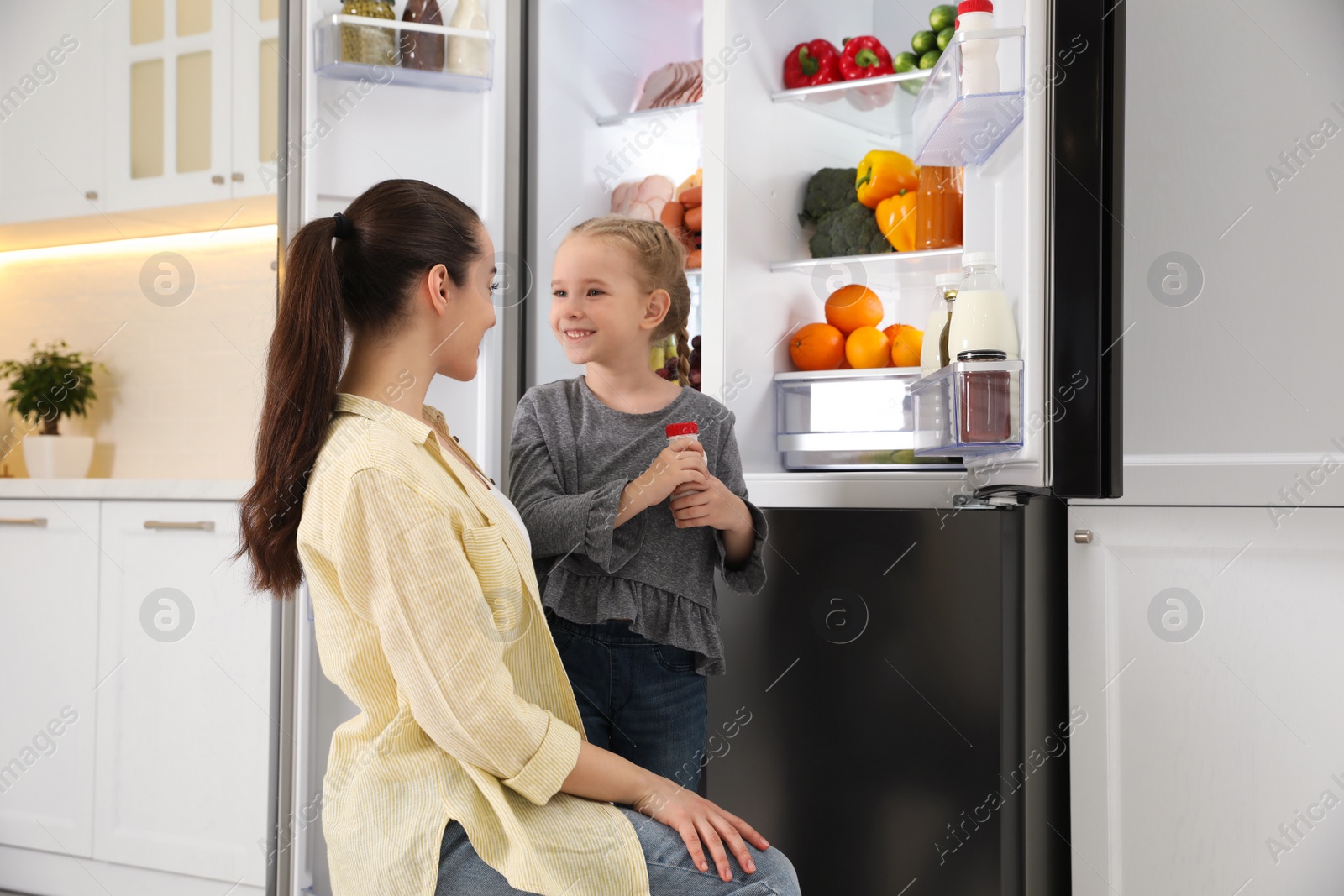 Photo of Young mother with daughter near open refrigerator in kitchen