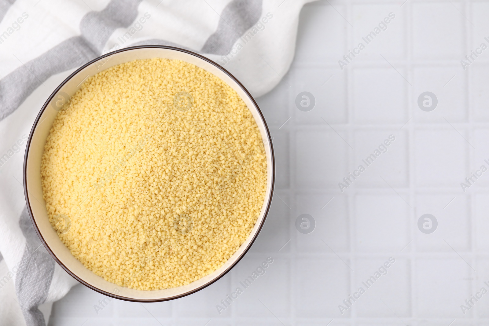 Photo of Raw couscous in bowl on white tiled table, top view. Space for text