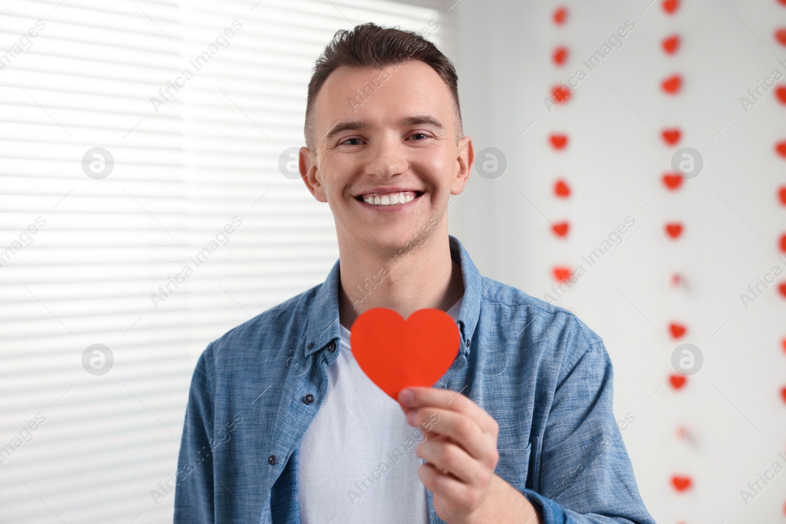 Photo of Handsome young man with decorative heart indoors, view from camera. Valentine's day celebration in long distance relationship