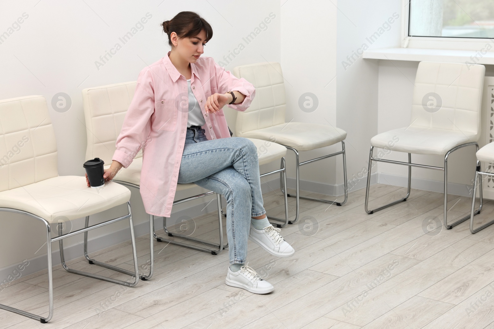 Photo of Woman with cup of drink looking at wrist watch and waiting for appointment indoors