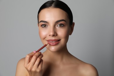 Pretty young woman applying beautiful nude lip pencil on grey background