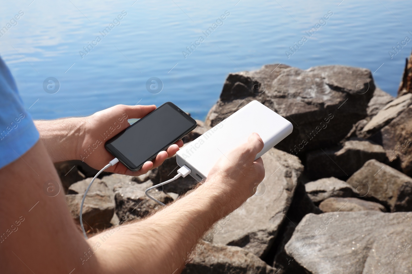 Photo of Man charging mobile phone with power bank on rocky mountain near river, closeup