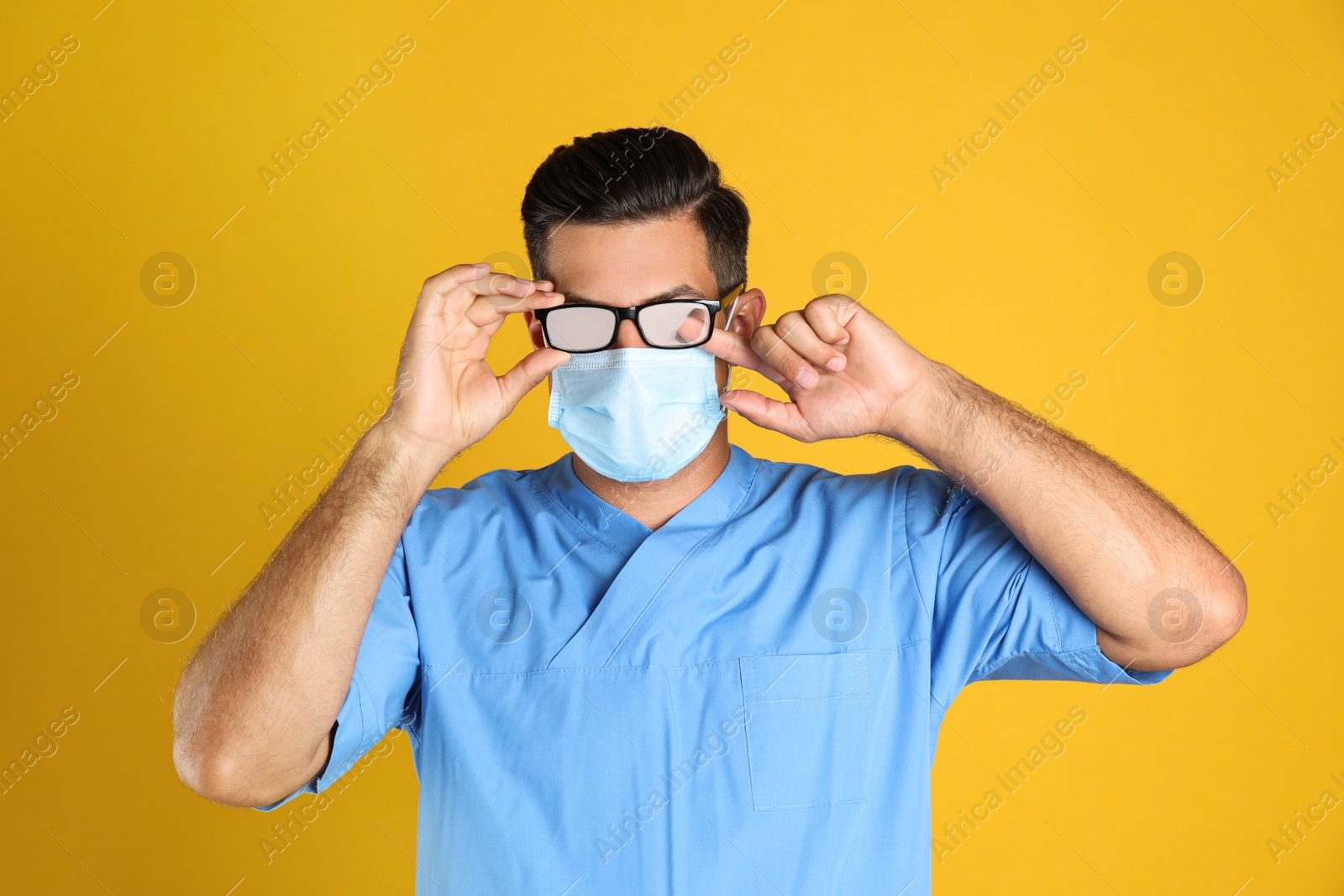 Photo of Doctor wiping foggy glasses caused by wearing medical mask on yellow background