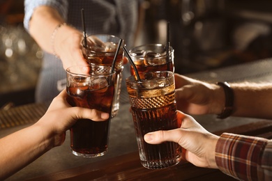 Photo of Group of friends clinking glasses with cola at table indoors, closeup