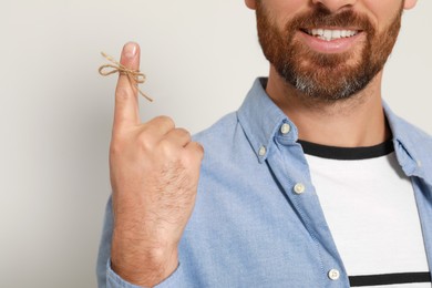 Smiling man showing index finger with tied bow as reminder on light grey background, closeup