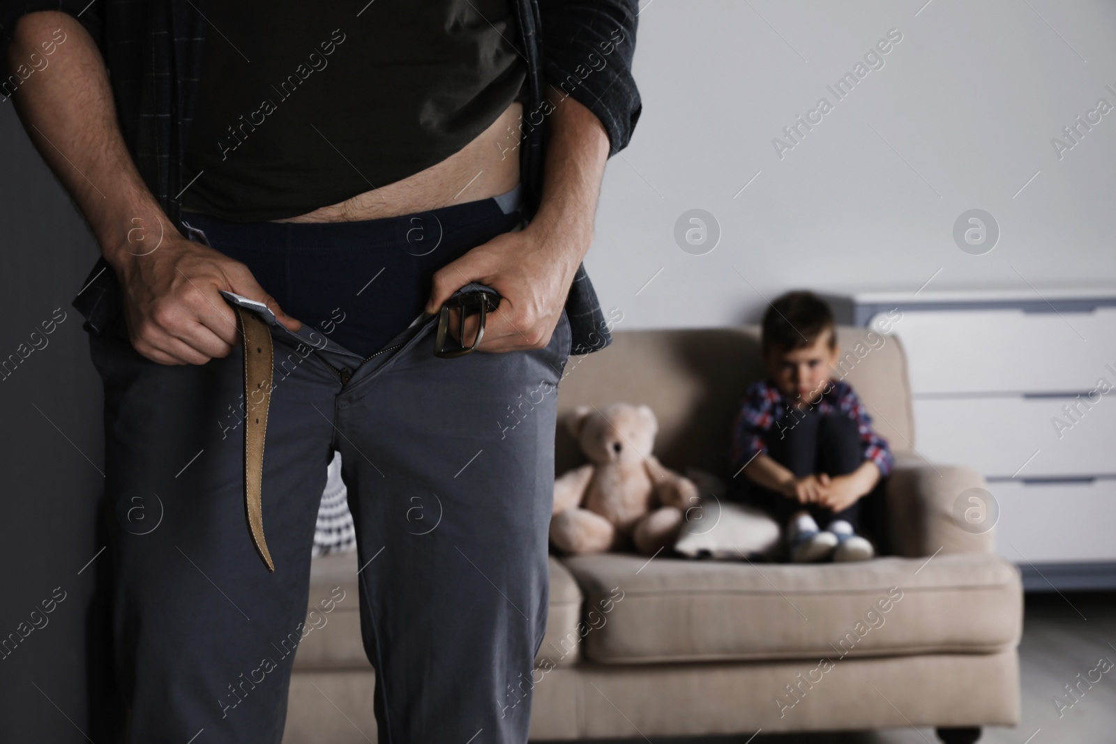 Photo of Man with unzipped pants standing near scared little boy on sofa indoors. Child in danger