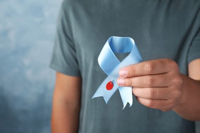 Man holding light blue ribbon with paper blood drop on color background, closeup. World Diabetes Day