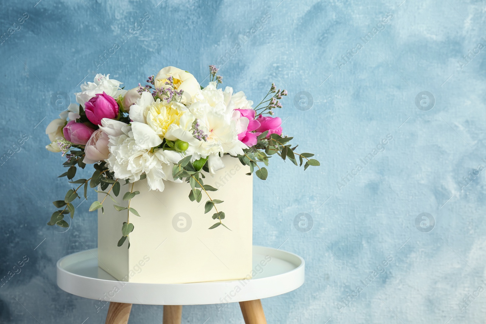 Photo of Box with bouquet of beautiful flowers on table against color background