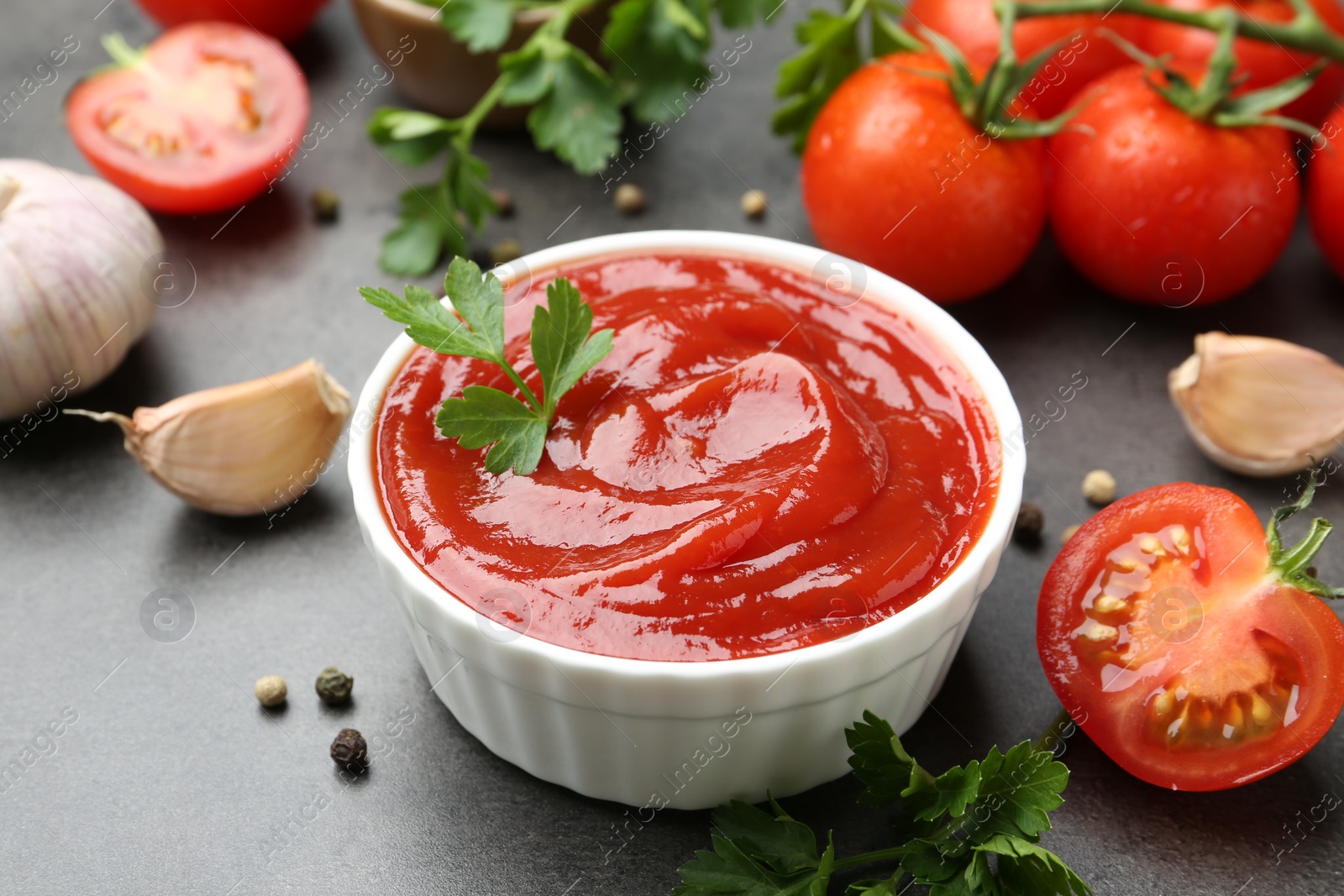 Photo of Delicious ketchup in bowl, tomatoes, parsley and garlic on grey table, closeup