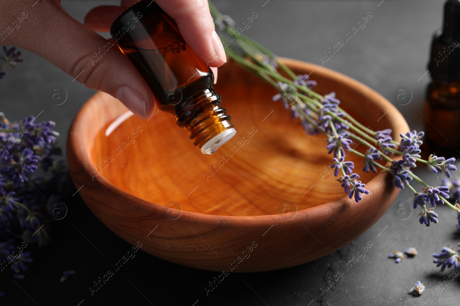 Photo of Woman dripping essential oil from bottle into bowl near lavender at grey table, closeup