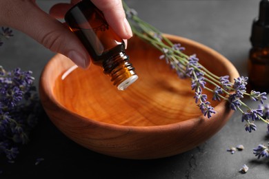 Woman dripping essential oil from bottle into bowl near lavender at grey table, closeup