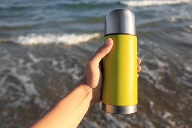Woman holding yellow thermos with hot drink on beach near sea, closeup. Space for text