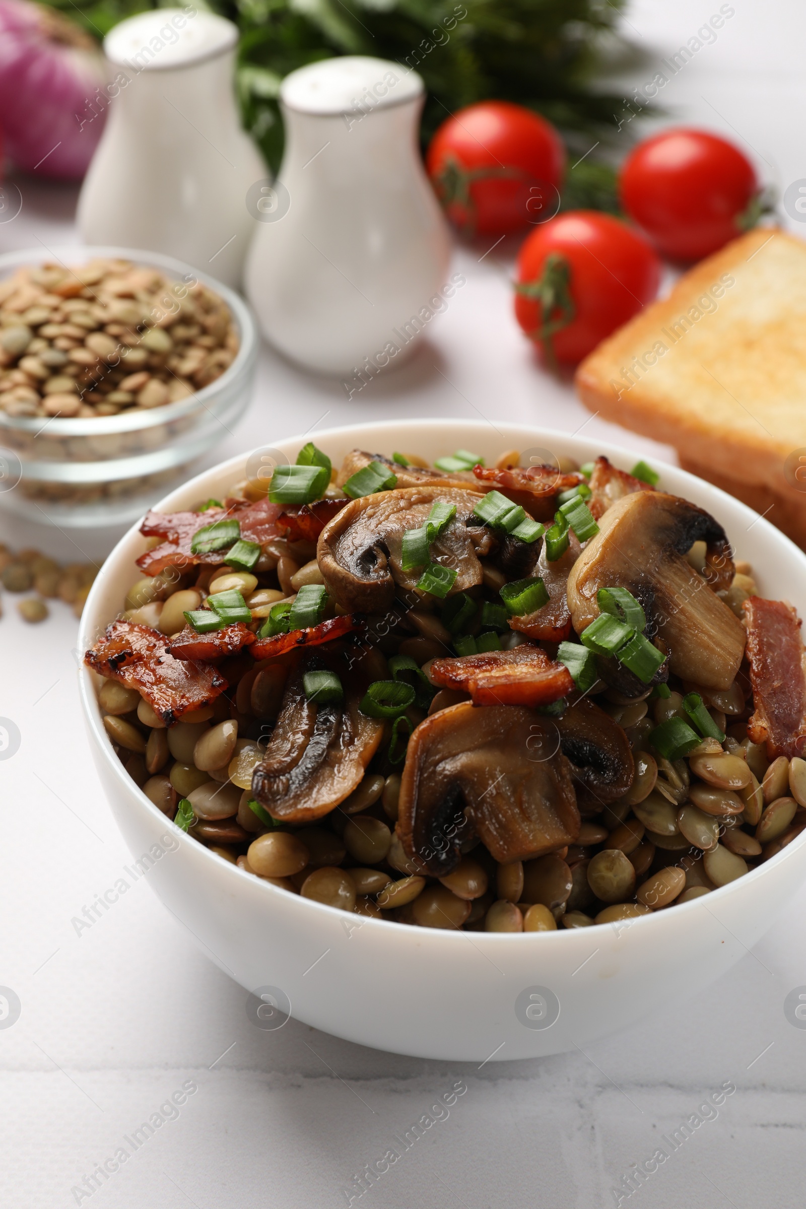 Photo of Delicious lentils with mushrooms, bacon and green onion in bowl served on white table, closeup
