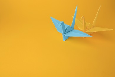 Photo of Origami art. Beautiful light blue and yellow paper cranes on orange background, space for text