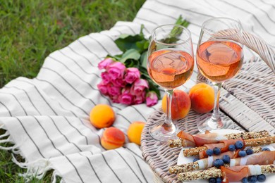 Photo of Glasses of delicious rose wine, food, flowers and basket on picnic blanket outdoors