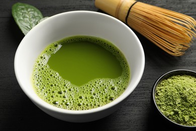 Photo of Cup of fresh matcha tea, green powder and bamboo whisk on black wooden table, closeup