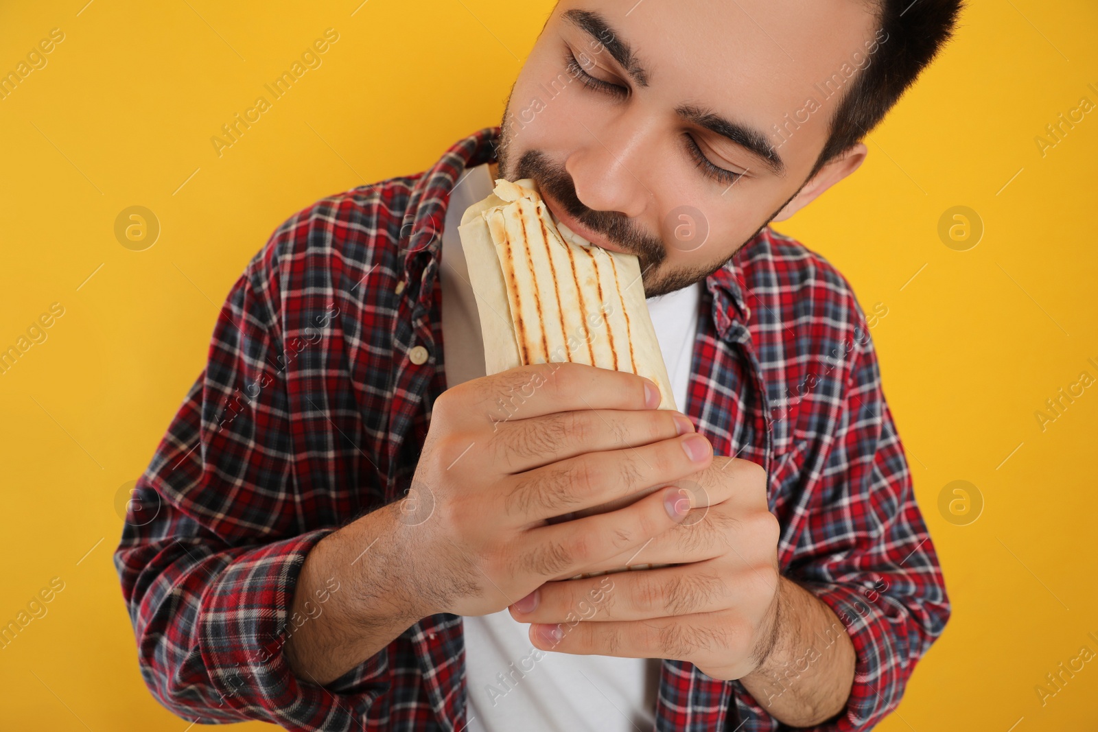 Photo of Young man eating tasty shawarma on yellow background, closeup