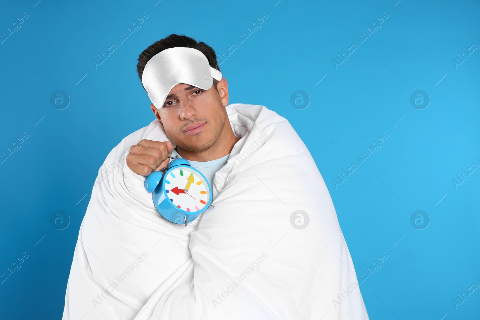 Photo of Man in sleeping mask wrapped with blanket holding alarm clock on blue background