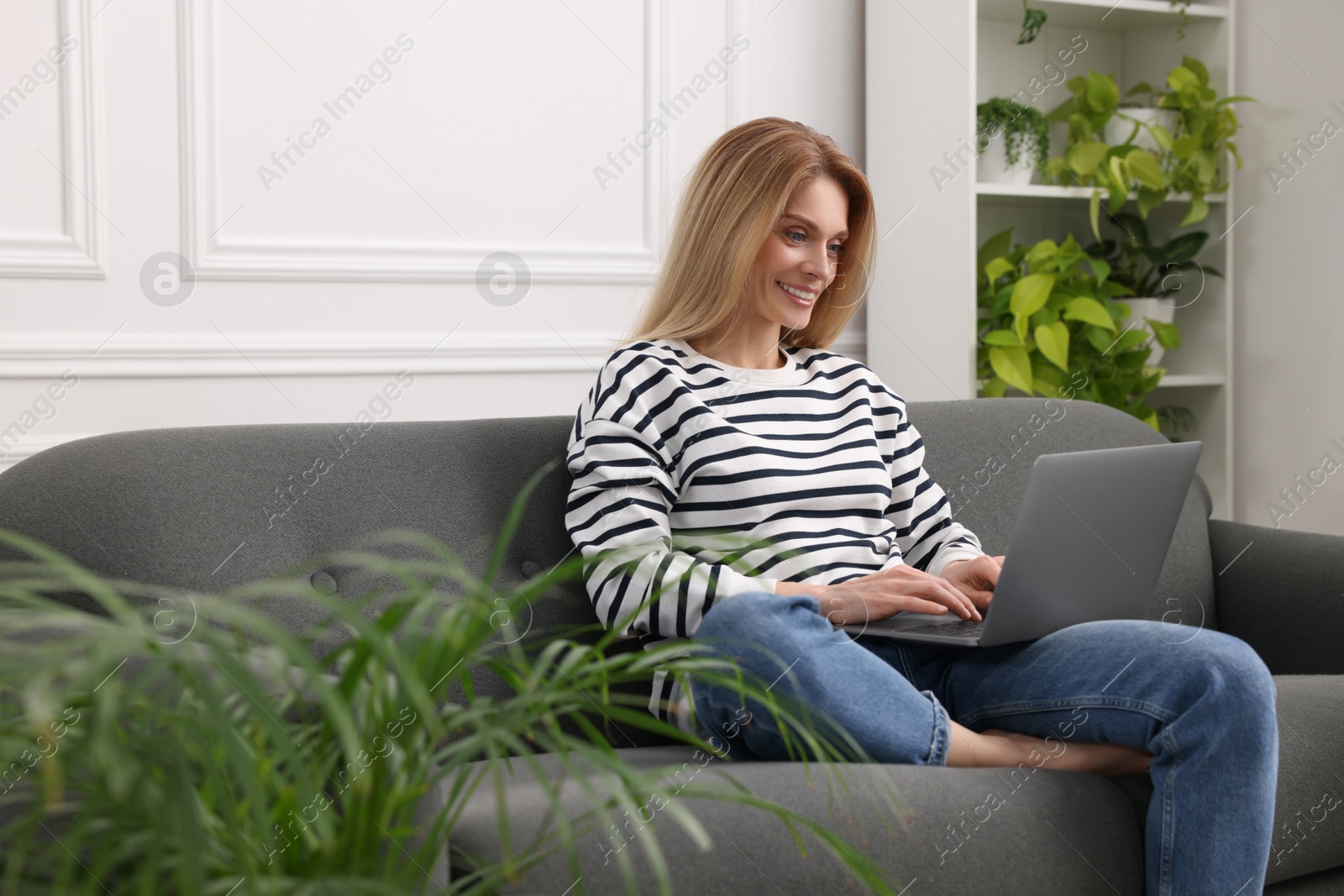 Photo of Woman working with laptop on sofa surrounded by beautiful potted houseplants at home