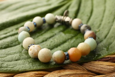 Photo of Beautiful bracelet with gemstones and green leaf on wicker surface, closeup