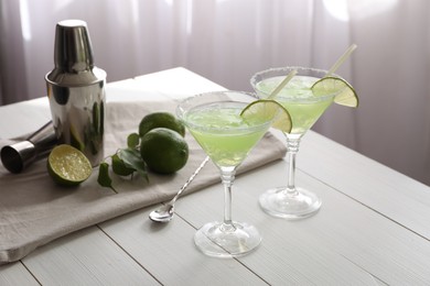 Photo of Delicious Margarita cocktail in glasses, limes and bartender equipment on white wooden table