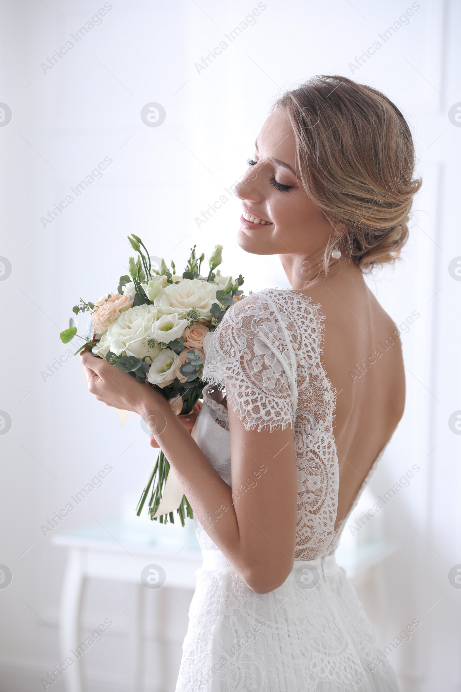 Photo of Young bride with elegant hairstyle holding wedding bouquet indoors