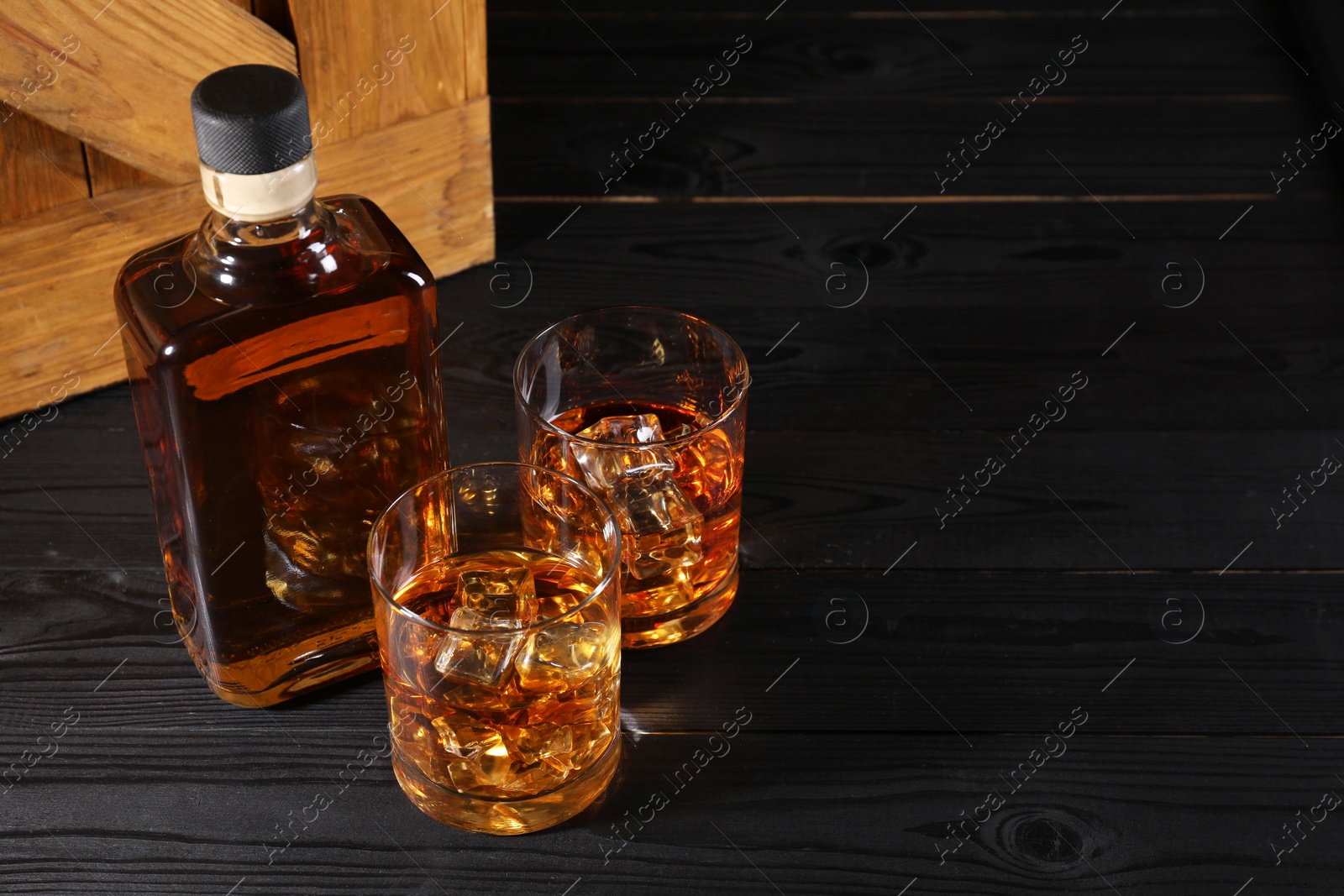 Photo of Whiskey with ice cubes in glasses and bottle on black wooden table, space for text