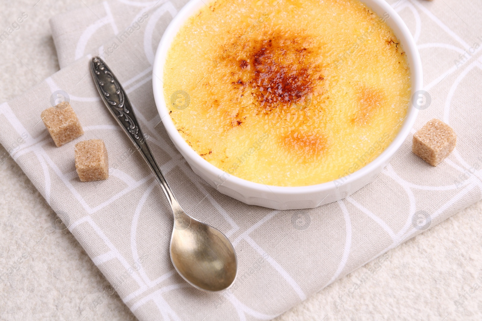 Photo of Delicious creme brulee in bowl, sugar cubes and spoon on light textured table, above view