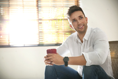 Young man with cup of drink relaxing near window at home. Space for text