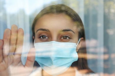 Stressed woman in protective mask looking out of window, view through glass. Self-isolation during COVID-19 pandemic