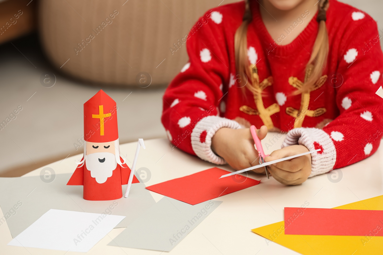 Photo of Cute little girl cutting paper at table with Saint Nicholas toy indoors, closeup