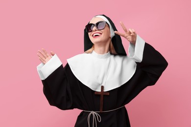 Happy woman in nun habit and sunglasses showing V-sign against pink background