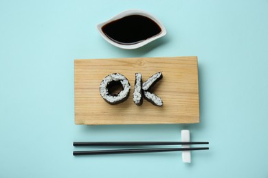 Photo of Word Ok made of sushi rolls, chopsticks and soy sauce on light turquoise background, flat lay