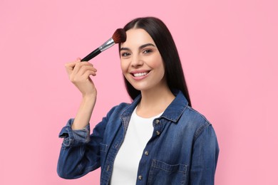 Beautiful woman applying makeup with brush on pink background