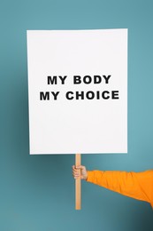 Image of Woman holding placard with phrase My Body My Choice on light blue background, closeup. Abortion protest