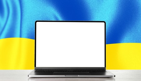 Image of Laptop with blank screen on table and Ukrainian national flag on background, space for text