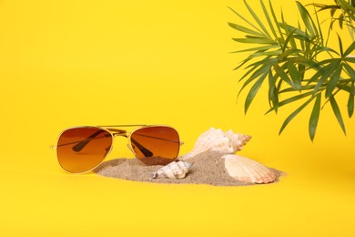 Stylish sunglasses, seashells, sand and palm leaves on yellow background