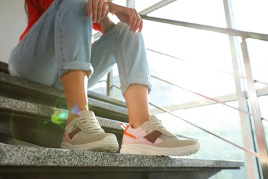 Photo of Young woman in stylish sneakers sitting on grey stairs indoors, closeup