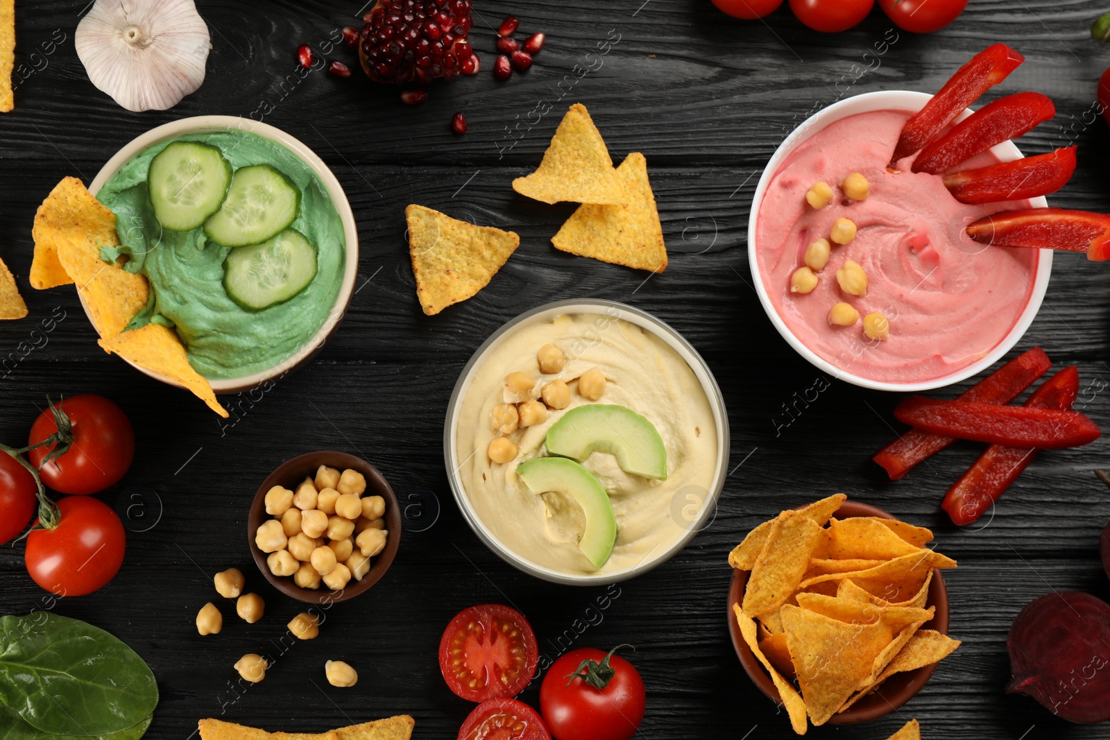 Photo of Different kinds of tasty hummus served with nachos on black wooden table, flat lay