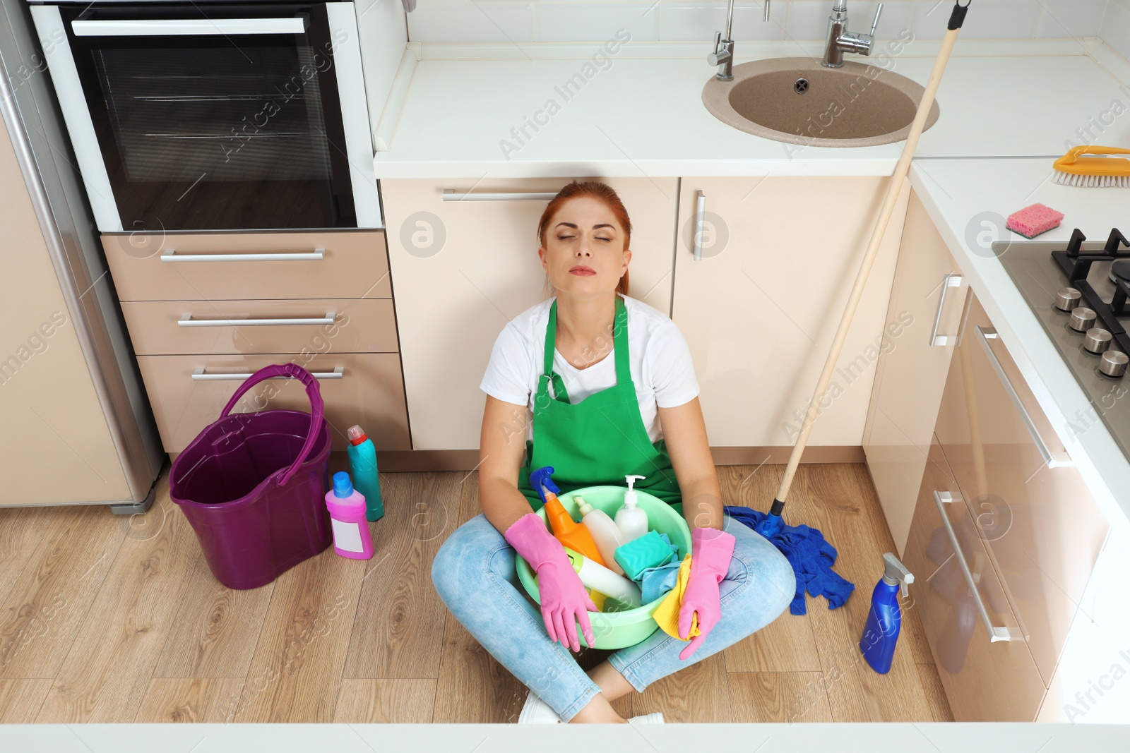 Photo of Tired woman with cleaning products sitting on floor in kitchen