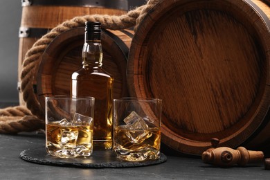 Photo of Whiskey with ice cubes in glasses, bottle and wooden barrels on black table