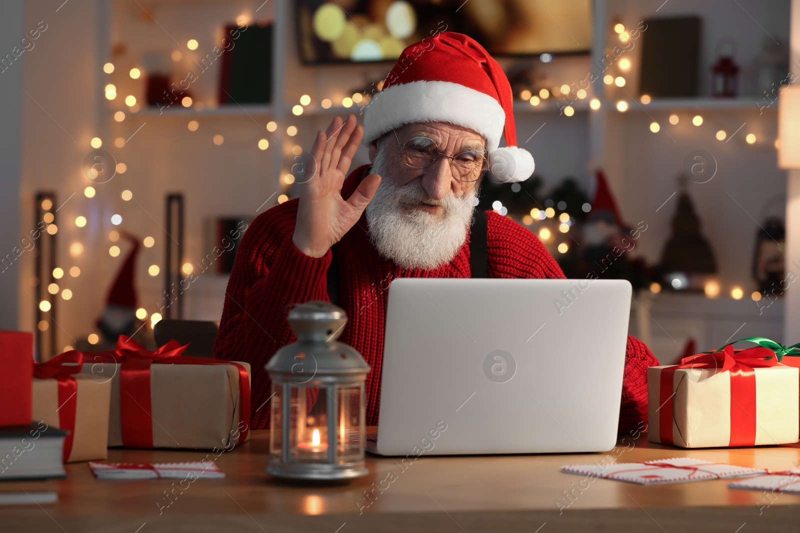 Photo of Santa Claus having video chat and waving hello at his workplace in room decorated for Christmas