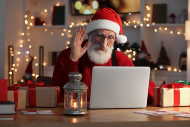 Santa Claus having video chat and waving hello at his workplace in room decorated for Christmas