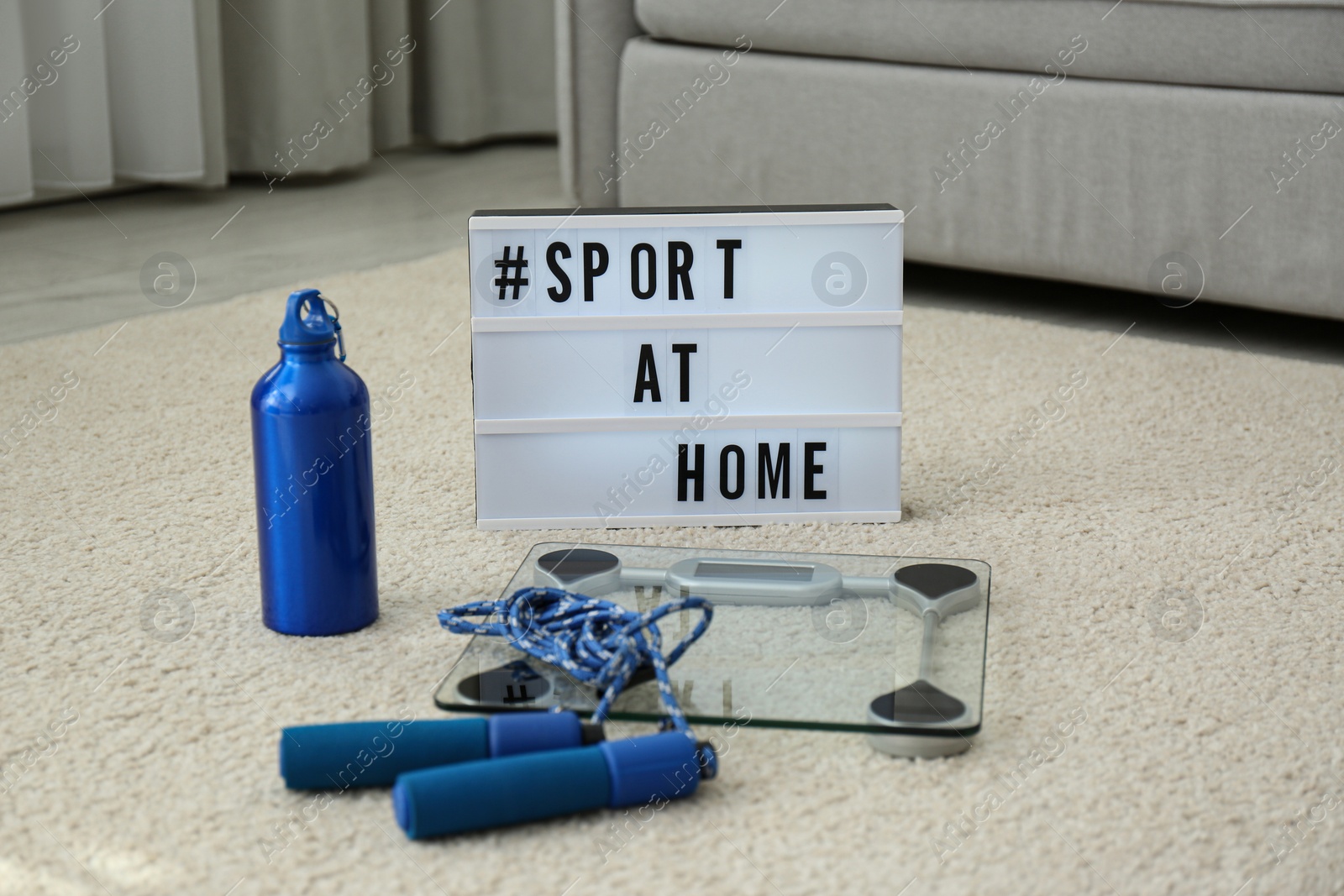Photo of Fitness equipment and lightbox with hashtag SPORT AT HOME on floor indoors. Message to promote self-isolation during COVID‑19 pandemic