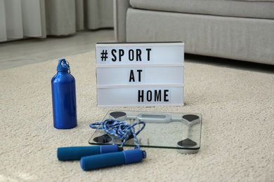 Fitness equipment and lightbox with hashtag SPORT AT HOME on floor indoors. Message to promote self-isolation during COVID‑19 pandemic